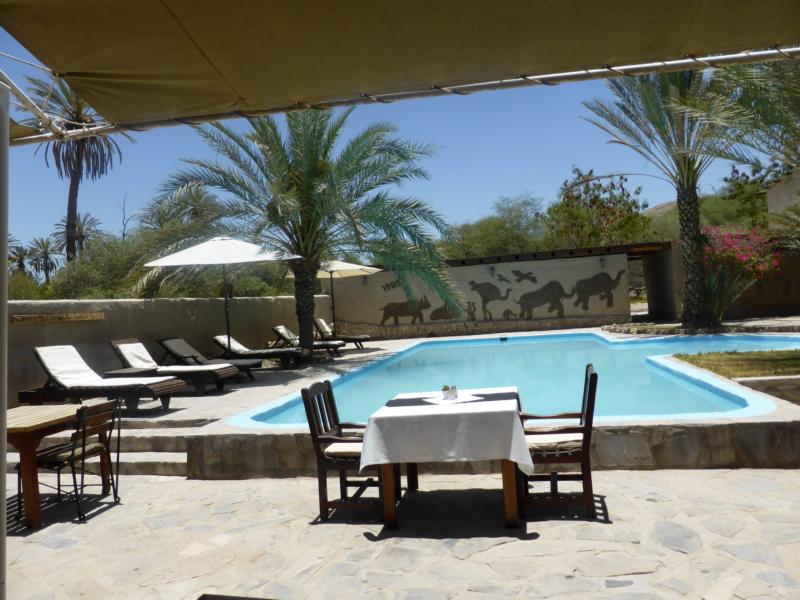 Fort Sesfontein: Auf der Sonnenterrasse vor dem Pool können die Mahlzeiten im Freien eingenommen werden Fort Sesfontein, Kaokoveld, Namibia, Trockenfluss, Gedeckter Tisch, Innenhof, Palmen, Pool, Sonnenliegen, Sonnenschirme, Afrikarma, Afrikarma Safaris, Afrikarma Safaris – Wildnis. Hautnah., afrikarma.de, Hoanib