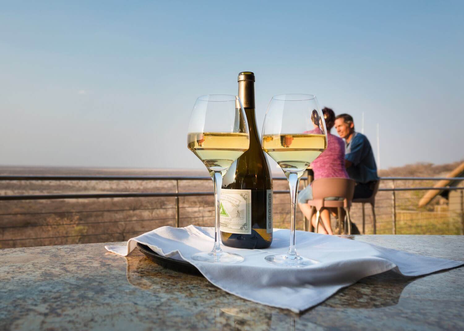 Afrikarma, Afrikarma Safaris, Afrikarma Safaris - Wildnis. Hautnah., Afrikarma.de, Ausblick auf die Savannenlandschaft, Serviette auf dem Tablet, Tisch, Weinflasche, Zaun, Zwei Gäste, Zwei Weingläser mit Wein, Safarihoek Lodge