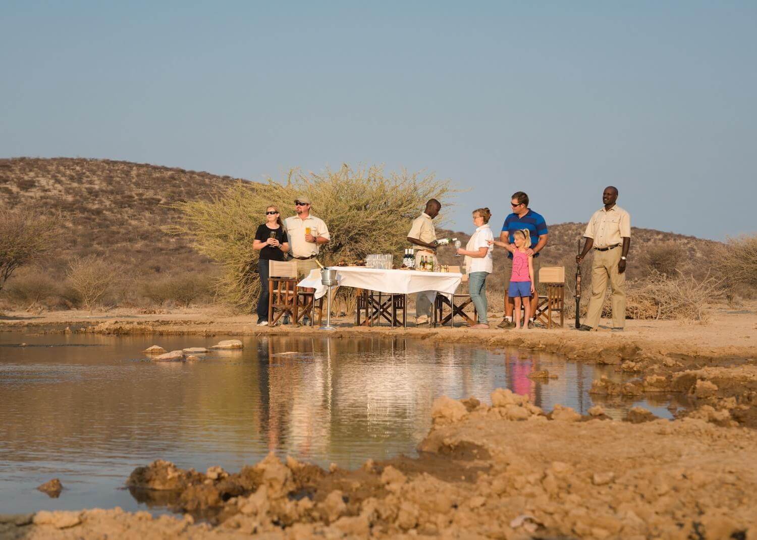 Afrikarma, Afrikarma Safaris, Afrikarma Safaris - Wildnis. Hautnah., Afrikarma.de, Büsche, Campingstühle, Gäste, Gedeckter Tisch, Ranger mit Gewehr, Wasserloch, Weinflaschen, Safarihoek Lodge