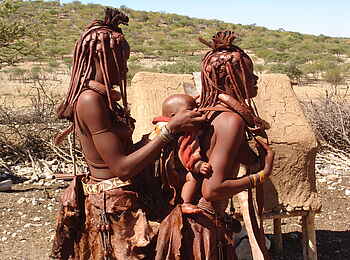 Fort Sesfontein: Eine Himba-Frau hilft einer zweiten ihr Baby auf dem Rücken zu tragen