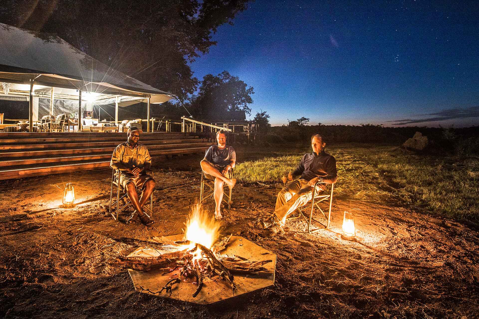 Afrikarma, Afrikarma Safaris, Afrikarma Safaris - Wildnis. Hautnah., Afrikarma.de, Deteema Springs Camp, Bäume, Büsche, Campingstühle, Gäste, Graslandschaft, Hauptbereich, Lagerfeuer, Paraffinlampen, Treppen