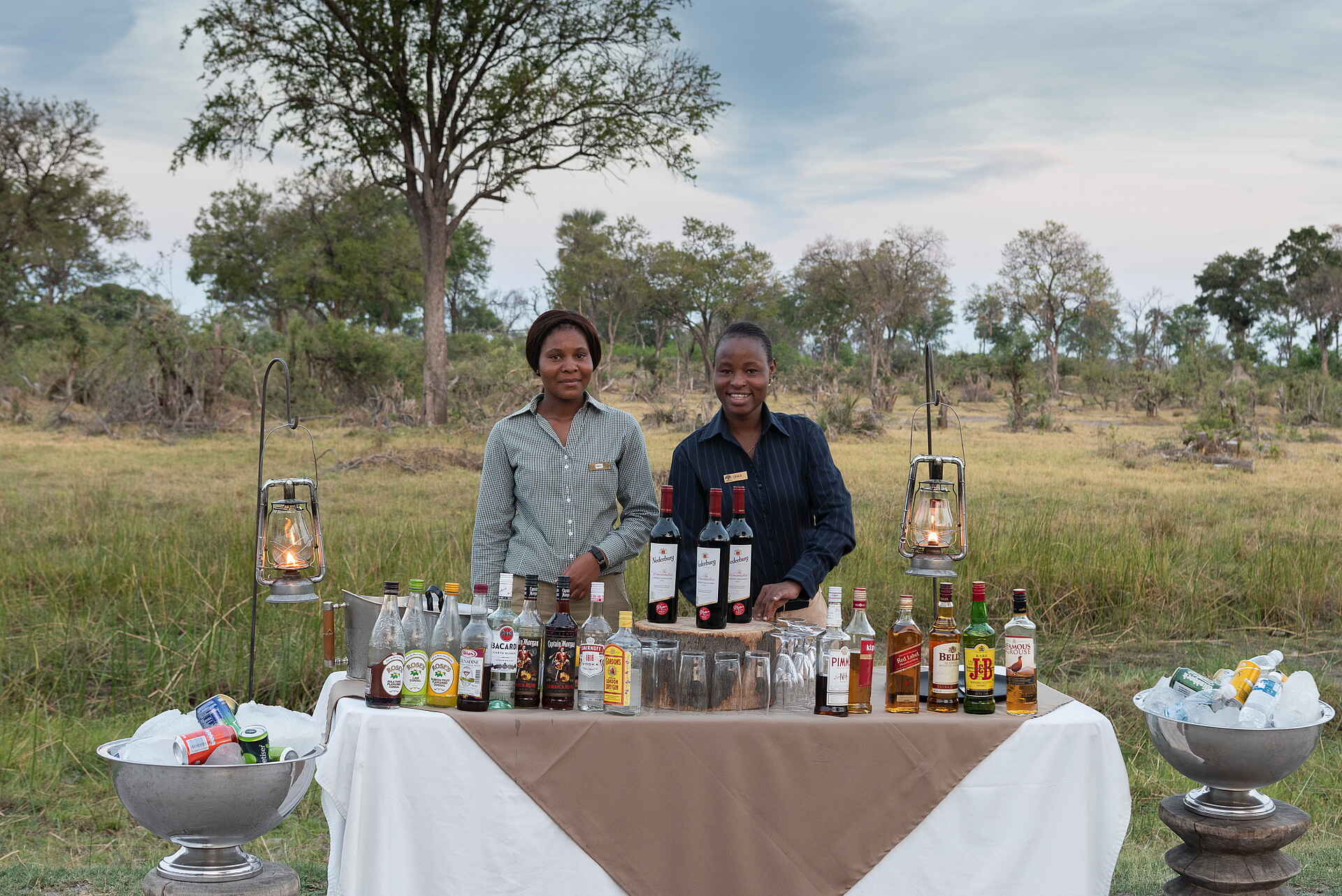 Gomoti Private Camp: Bar im Außenbereich Afrikarma, Afrikarma Safaris, Afrikarma Safaris. Wildnis. Hautnah., Afrikarma.de, Camp, Gomoti Private, Gomoti Private Camp, Bar, Bäume, draußen, Eiswürfel, Lampen, Personal, Tischdecke, Wein