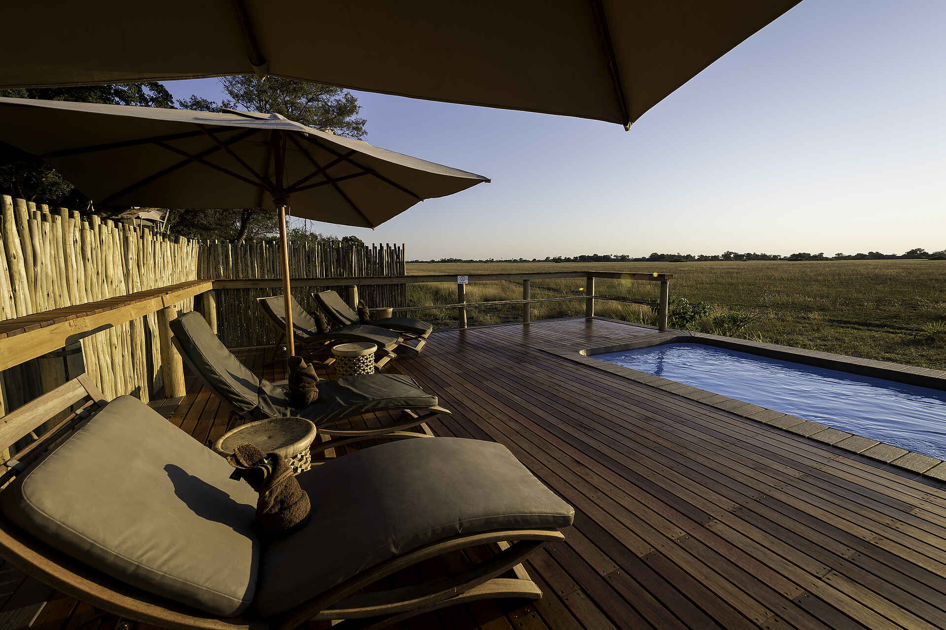 Kwetsani Camp: Swimmingpool Afrikarma, Afrikarma Safaris, Afrikarma Safaris - Wildnis. Hautnah., Afrikarma.de, Kwetsani Camp, Baumkette, Holzdeck, Holzzaun, Liegestühle, runde Holztische, Savannenlandschaft, Sonnenschirme, Swimmingpool