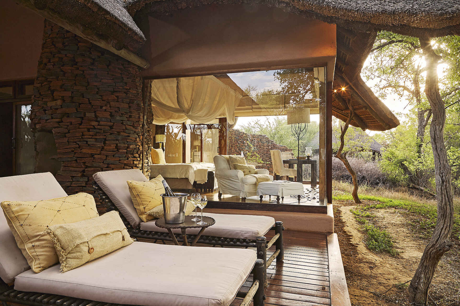 Afrikarma, Afrikarma Safaris, Afrikarma Safaris - Wildnis. Hautnah., Afrikarma.de, Bäume, Büsche, Holzdeck, Kissen, Liegestühle, Sanctuary Makanyane Luxury Safari Lodge, Sofasitze, Strohdach, Tische, Weinflasche, Weingläser, Weinkühler, Bett, Decken, Lampen