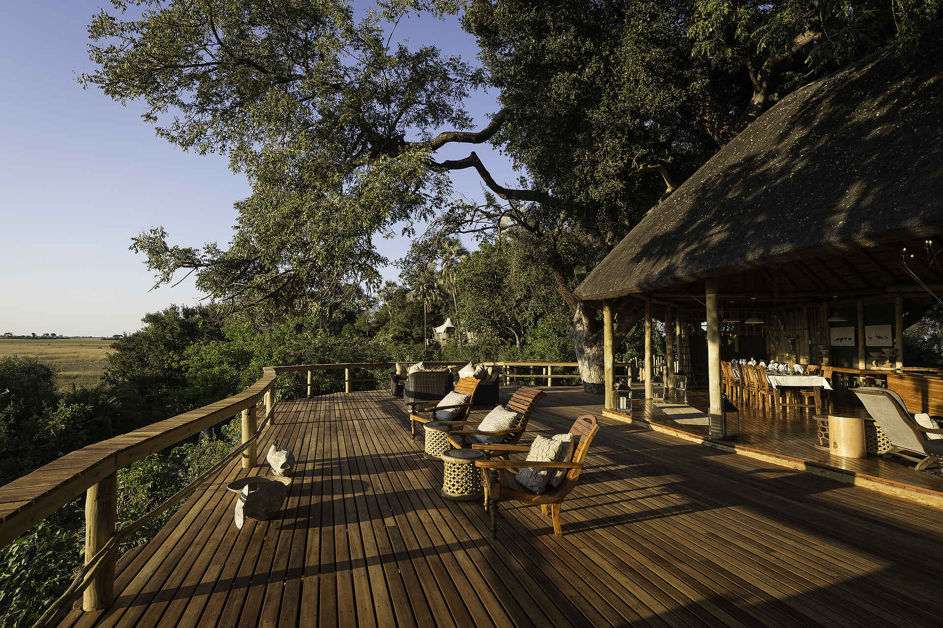 Kwetsani Camp: Außendeck der Lodge Afrikarma, Afrikarma Safaris, Afrikarma Safaris - Wildnis. Hautnah., Afrikarma.de, Kwetsani Camp, Außendeck der Lodge, Bäume, Esszimmertisch, Kissen, runde Holztische, Strohdach, Stühle