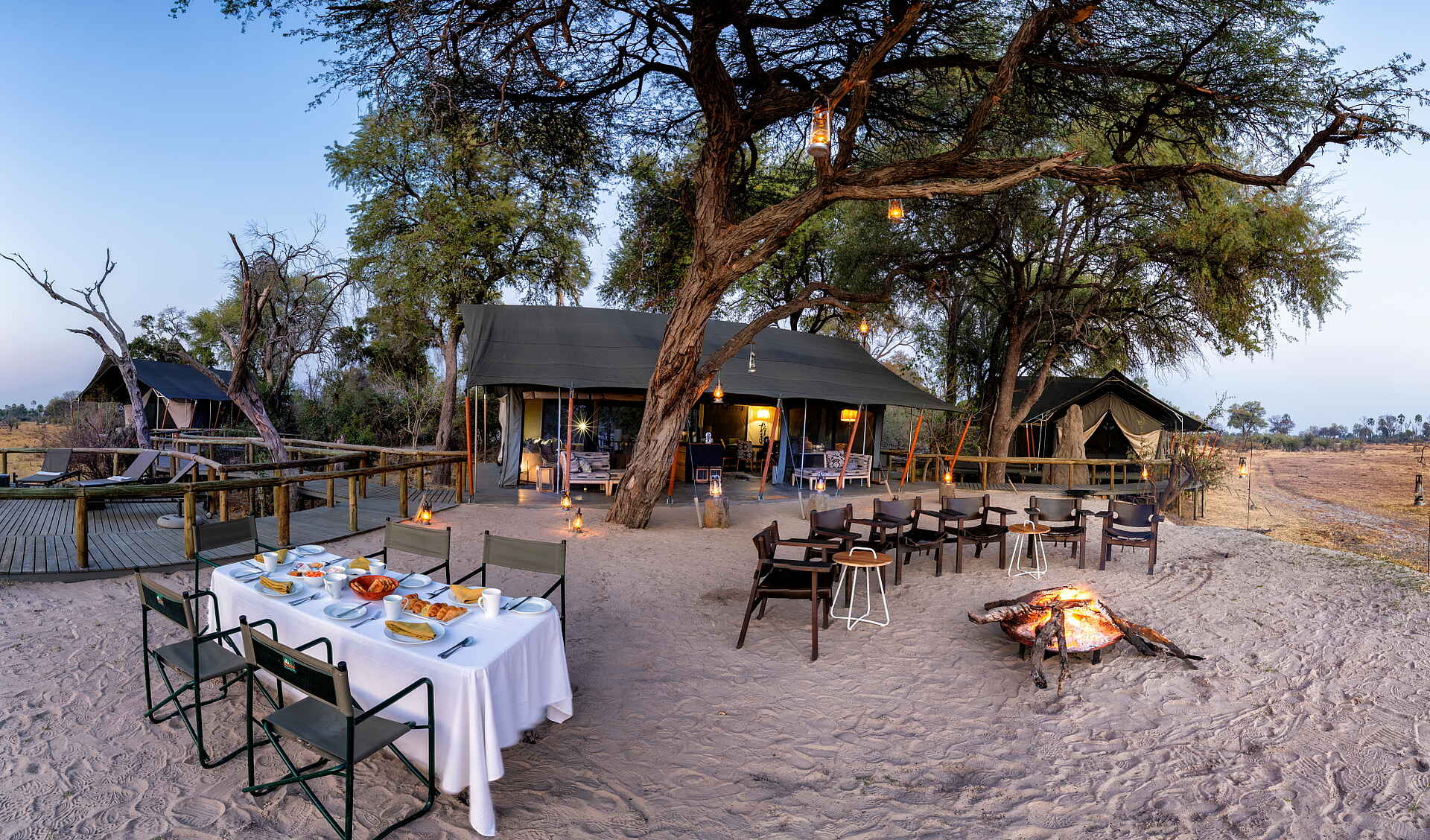 Gomoti Private Camp: Feuerstelle und Essbereich Afrikarma, Afrikarma Safaris, Afrikarma Safaris. Wildnis. Hautnah., Afrikarma.de, Camp, Gomoti Private, Gomoti Private Camp, Bäume, draußen, Essstühle, Esstisch, Feuerschale, Feuerstelle, Holz, Lounge, Sand, Stühle