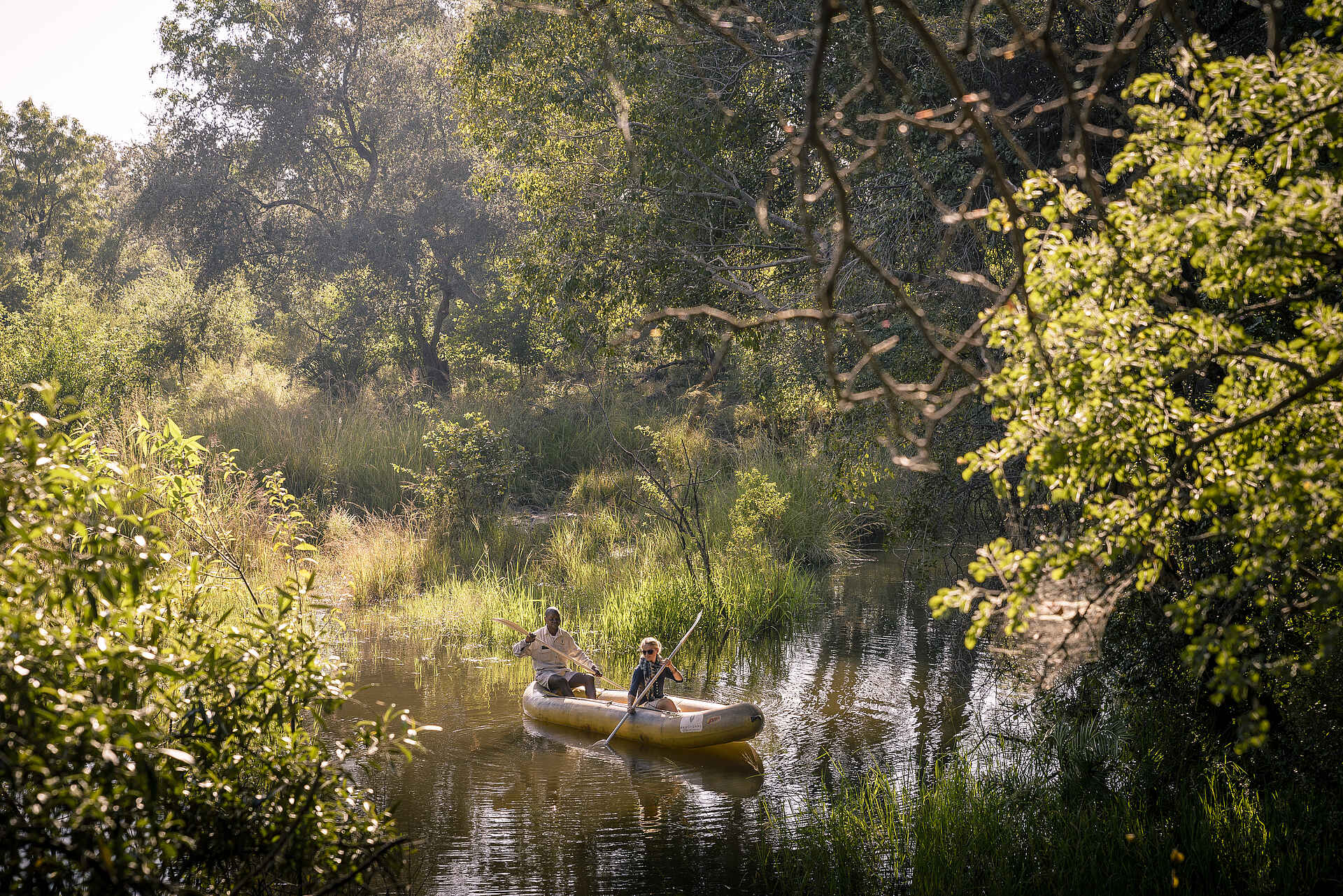 Sanctuary Sussi & Chuma: Kanufahrt auf dem Zambezi River Afrikarma, Afrikarma Safaris, Afrikarma Safaris - Wildnis. Hautnah., Afrikarma.de, Sanctuary Sussi & Chuma, Bäume, Büsche, Gast, Gras, Kanu, Kanufahrer, Wasser