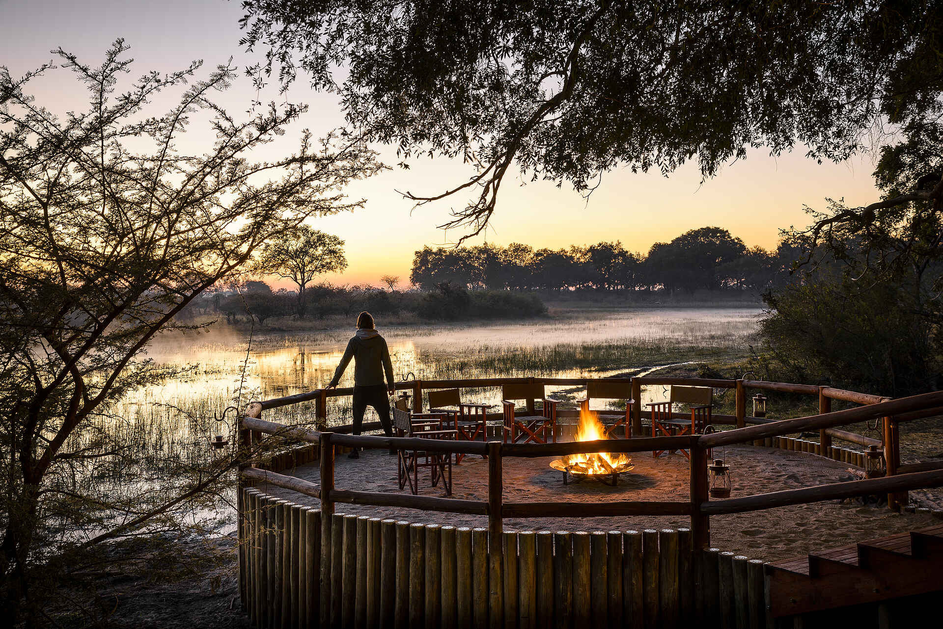 Afrikarma, Afrikarma Safaris, Afrikarma Safaris - Wildnis. Hautnah., Afrikarma.de, Sanctuary Chief's Camp, Bäume, Boma, Büsche, Campingstühle, Gast, Holzzaun, Lagerfeuer, Paraffinlampen, Sandboden, Savannenlandschaft