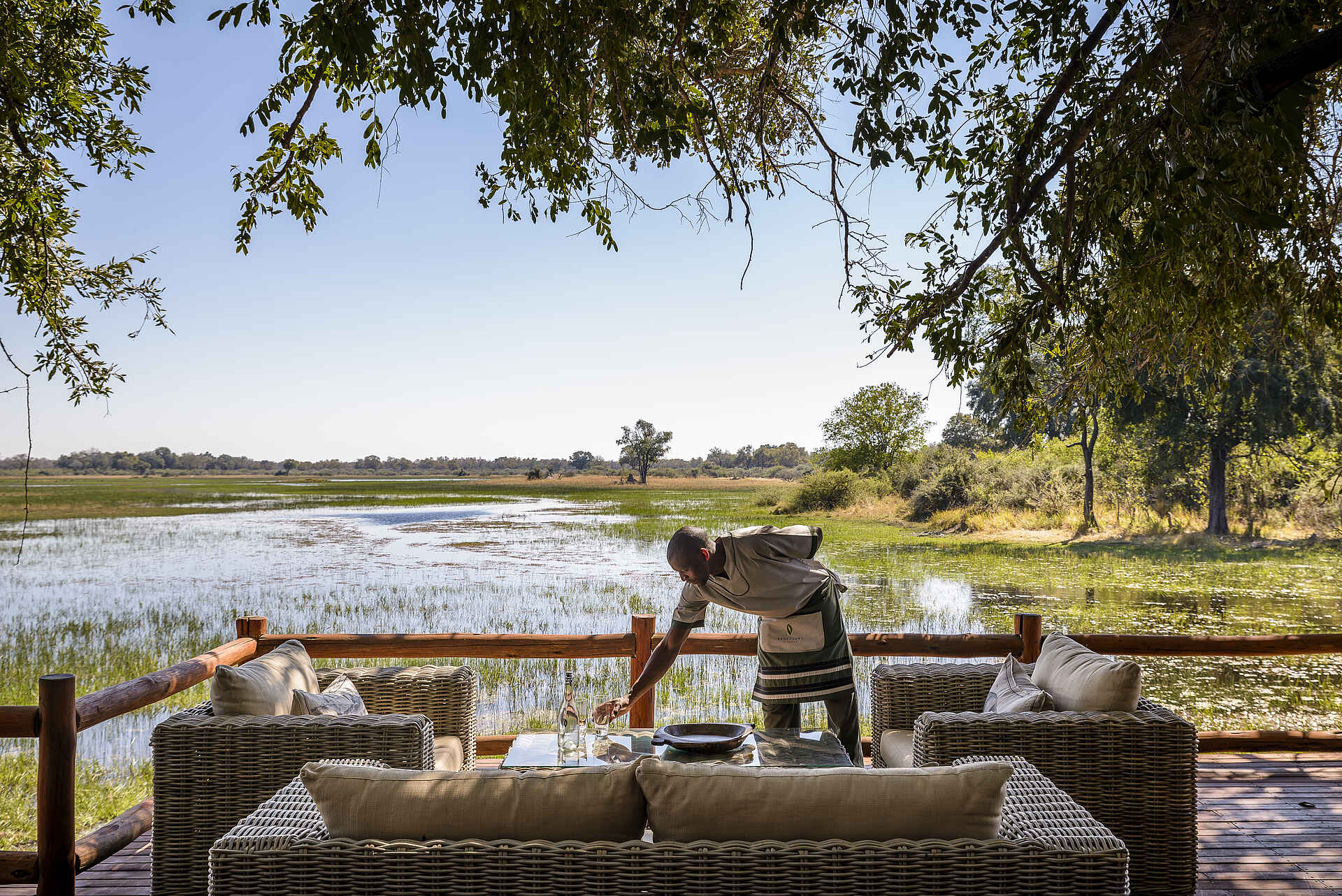 Afrikarma, Afrikarma Safaris, Afrikarma Safaris - Wildnis. Hautnah., Afrikarma.de, Sanctuary Chief's Camp, AnAngestellter, Bäume, Büsche, Flasche, Fluß, Gläser, Graslandschaft, Kissen, Ornamente, Sofasitze, Tisch