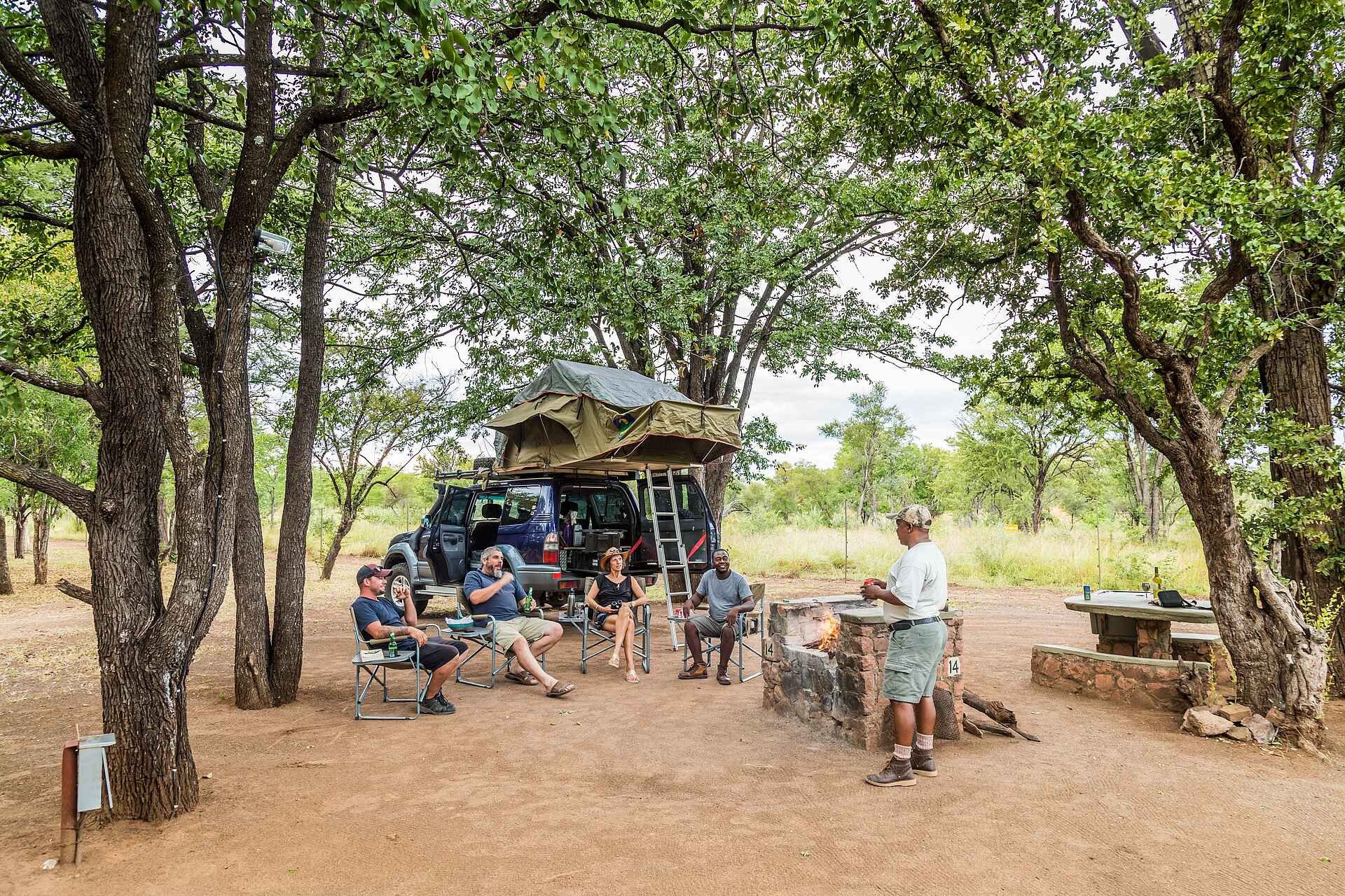 Afrikarma, Afrikarma Safaris, Afrikarma Safaris - Wildnis. Hautnah., Afrikarma.de, Robins Camp, Bäume, Campingstühle, Dachzelt, Fernglas, Feuer, Flaschen, Gäste, Gläser, Grillplatz, Landrover, Leiter, Steinbank, Tisch